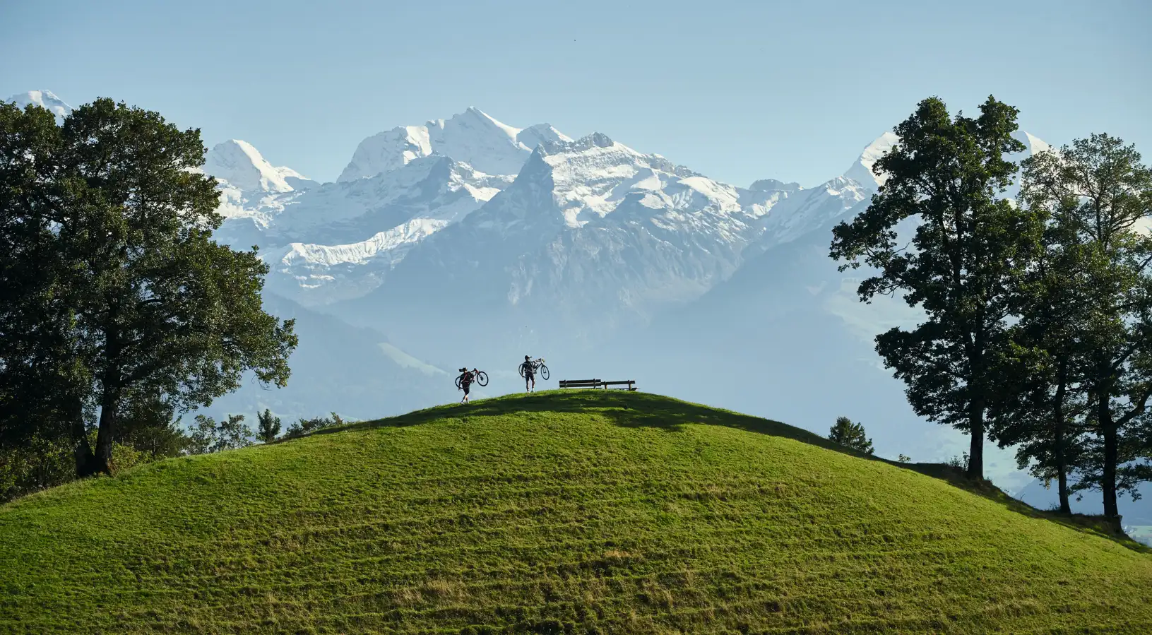 Two cyclists carrying their bikes over a high grassy ridge with a panoramic mountain backdrop, showcasing active outdoor things to do in Interlaken.